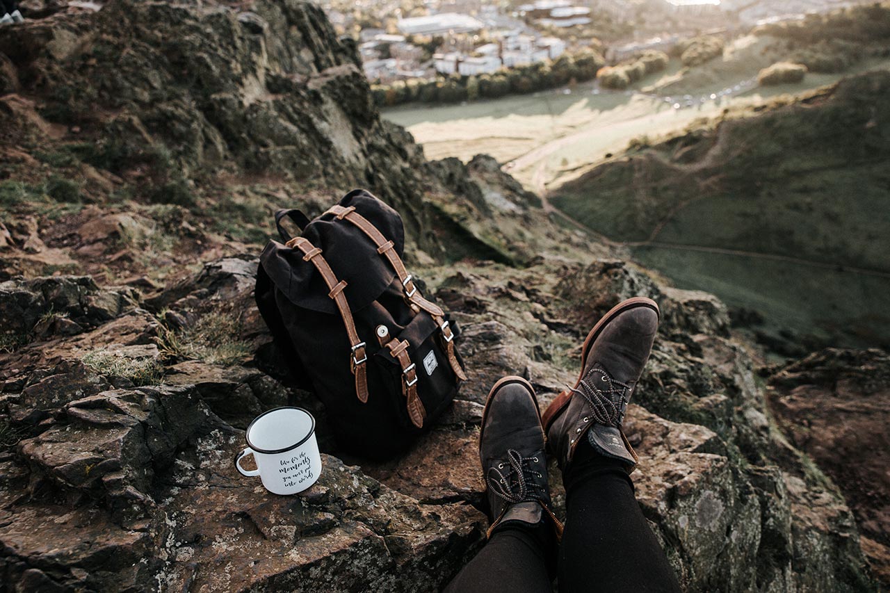 een rugzak met een kopje en voeten met wandelschoenen op een berg met uitzicht op het dal